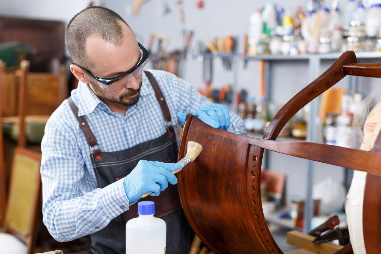 A man wearing an apron is focused on crafting a wooden chair in a workshop setting.