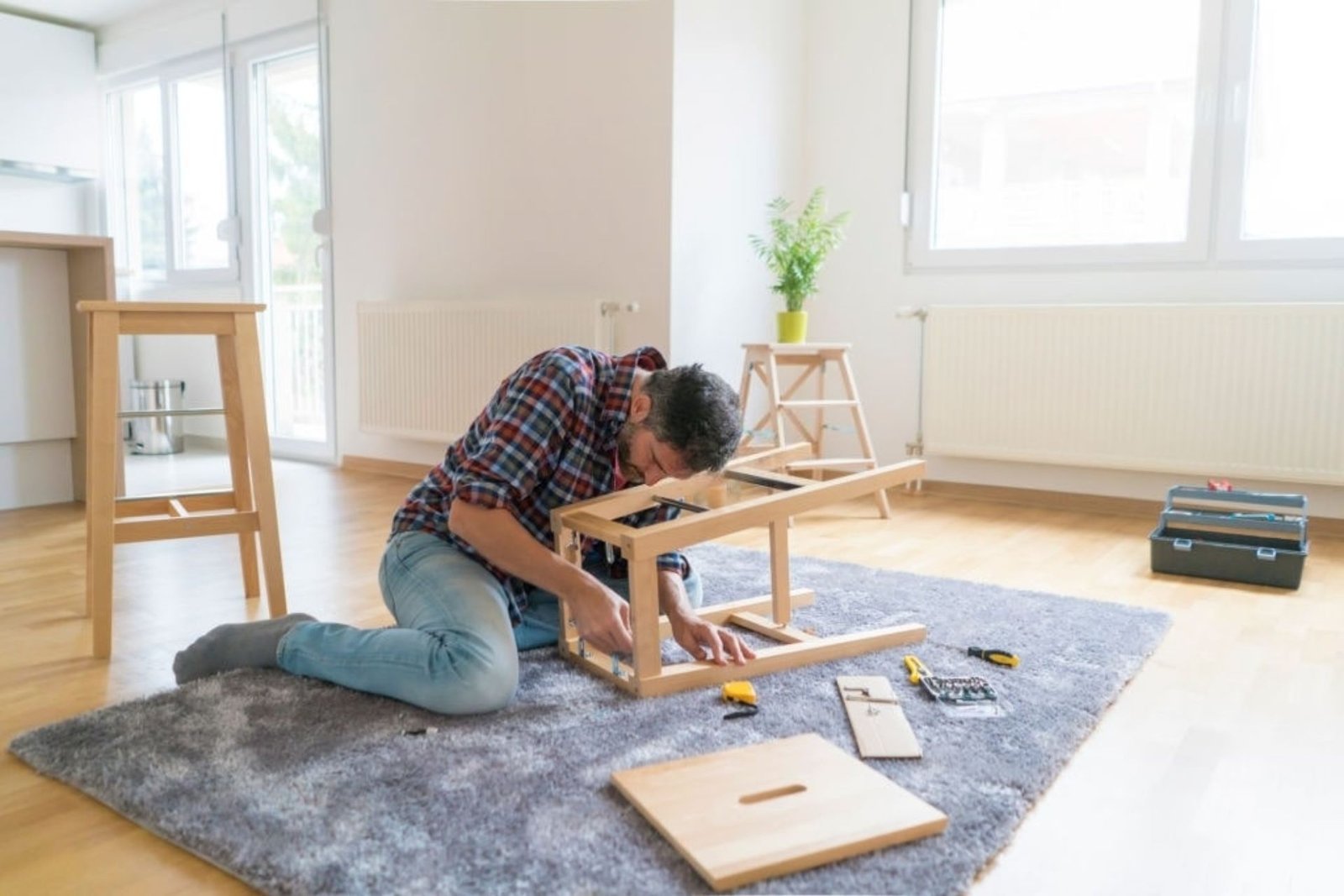 A man works diligently at a wooden table in his living room, surrounded by tools and personal items.