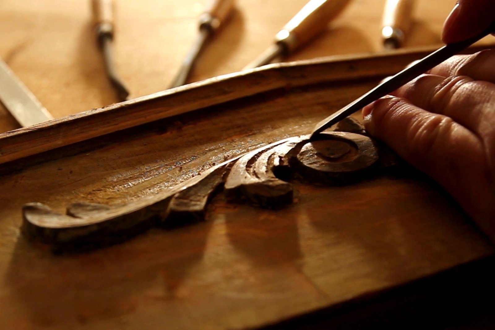 A person carves a wooden piece with a chisel, concentrating on the craftsmanship and wood texture.