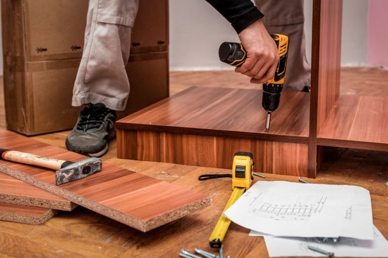 A man operates a drill while constructing a wooden floor for a furniture repair service.