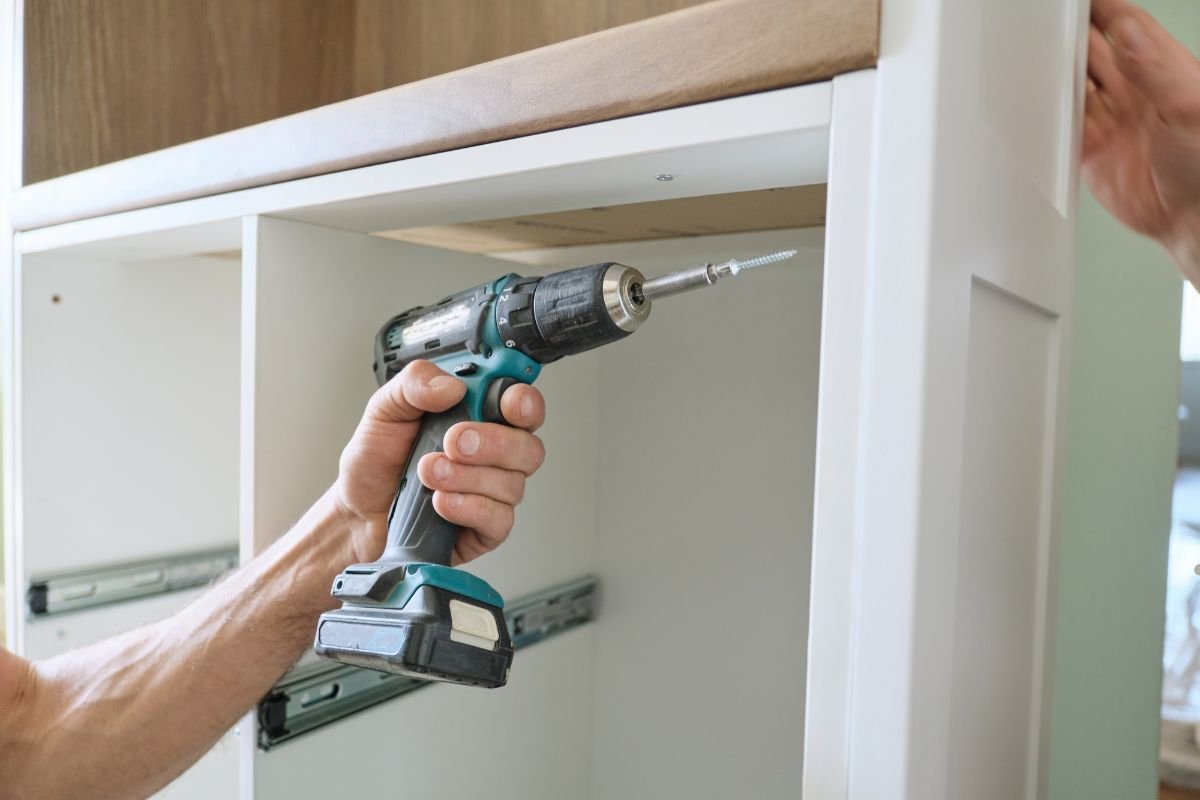 A man operates a drill to install a cabinet as part of a furniture repair service.