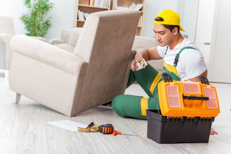 A man in a hard hat and overalls sits on the floor surrounded by tools, working on furniture repair.